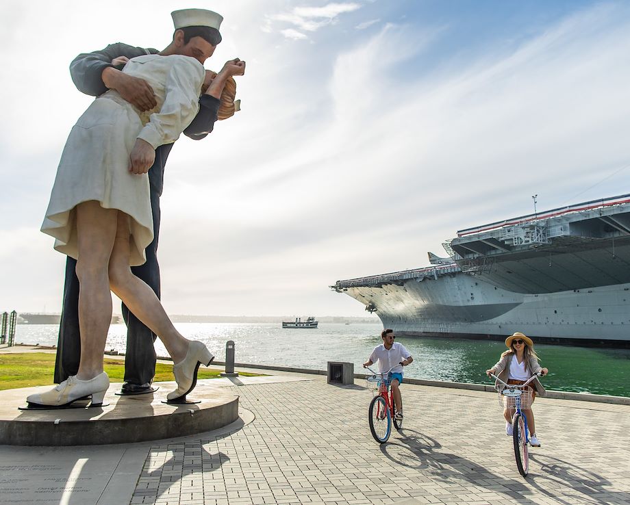 A man and woman bicycling next to a statue of a man and woman embracing. A naval ship is in the background.